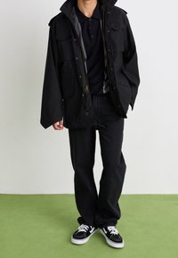 Czarna wodoodporna kurtka z wieloma kieszeniami, szara podszewka, w połączeniu z ciemnymi dżinsami i czarnymi trampkami na zielonej podłodze.