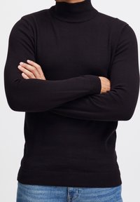 Pull à col roulé noir à manches longues, col et poignets côtelés, coupe ajustée, porté avec un jean en denim bleu. Texture lisse.