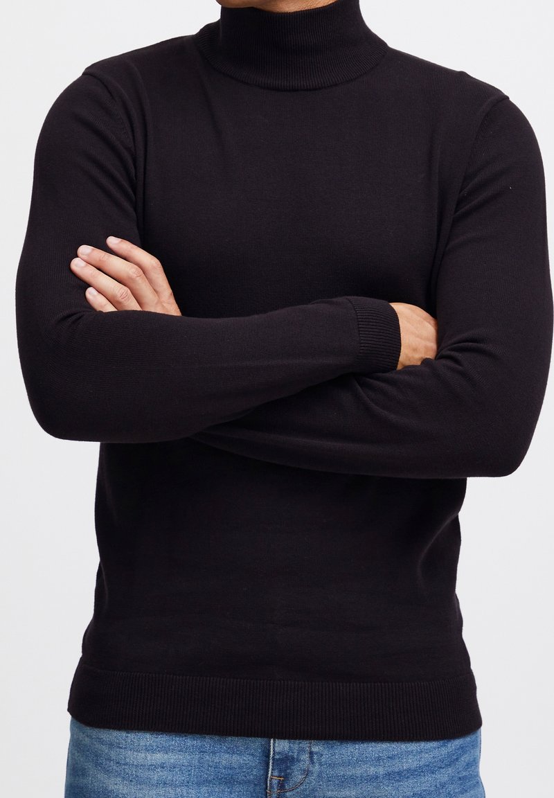 Pull à col roulé noir à manches longues, col et poignets côtelés, coupe ajustée, porté avec un jean en denim bleu. Texture lisse.