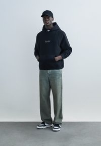 Man wearing black baseball cap and black hoodie with white text, faded loose jeans, and black-and-white sneakers, standing on gray floor.