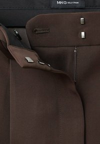 Pantalons en tissu marron avec une texture lisse, dotés d'une fermeture éclair et d'accents en métal argenté. L'étiquette intérieure indique "MNG Selection".