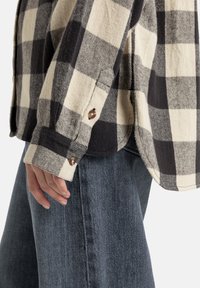 Flannel shirt with a large black and cream check pattern, featuring a buttoned cuff and a soft texture. Paired with dark denim pants.