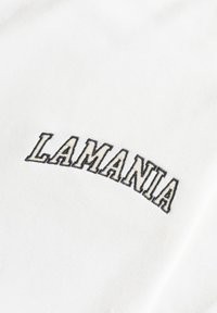 Bílá látka s nápisem "LAMANIA" v tučném, černým obrysem vyšitým písmem. Textura vypadá jemně a hladce, bez dalších vzorů nebo akcentů.