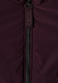 Veste zippée bordeaux avec une fermeture éclair noire et un tirant en tissu étiqueté "CECIL", dotée d'une texture lisse et légèrement brillante.