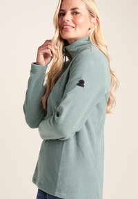 Blonde woman in light blue fleece jacket with high collar, smiling and touching her hair, standing against a plain background.