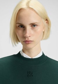 Felpa verde a coste con "HUGO" ricamato in nero. Il modello ha i capelli biondi corti e indossa una camicia bianca con colletto sotto.