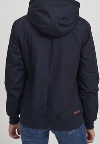 Granatowa kurtka z kapturem, ściągaczami na mankietach i pasie. Posiada skórzaną naszywkę z logo na dolnej stronie oraz gładką zewnętrzną tkaninę.