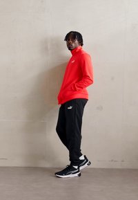 Rode lange mouw sporttop, zwarte broek en zwarte sneakers, met een strak ontwerp en zichtbare merknamen op de borst en pijp.