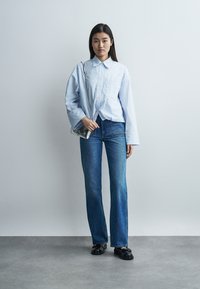 Γυναίκα στέκεται φορώντας μπλε και λευκό ριγέ φαρδιά πουκάμισα, μπλε τζιν, μαύρα παπούτσια και κρατώντας μια μικρή ασημένια τσάντα στον ώμο της.