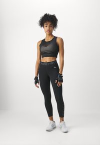 Črni športni krojeni top z mrežastimi detajli, v kombinaciji z visokimi črnimi leginami, na katerih je oznaka "NIKE PRO", rokavicami in belimi športnimi čevlji.