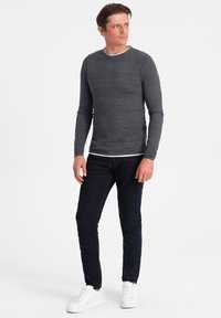 Graues Langarmshirt mit strukturiertem Design und weißem Rundhalsausschnitt, kombiniert mit dunklen Jeans und weißen Sneakern.