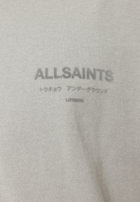 Lys grå T-shirt lavet af blødt, struktureret stof med præget "ALLSAINTS" branding og japansk tekst i subtil grå.