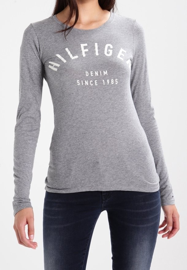 Kvinna som bär en figursydd grå långärmad tröja med texten "HILFIGER DENIM SINCE 1985" och mörkblåjeans.