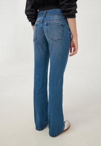 Niebieskie dżinsy z rozszerzanymi nogawkami i wysokim stanem, z dwoma tylnymi kieszeniami i widocznym szwem. Noszone z białymi butami i czarną bluzką.