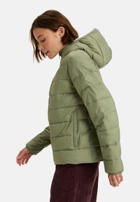 Chaqueta verde acolchada con capucha, que presenta secciones de acolchado horizontal y bolsillos laterales. Textura suave, material sintético ligero.