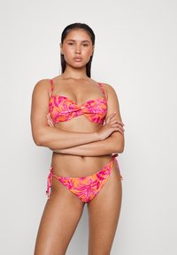 Conjunto de biquíni com um padrão floral tropical em tons de rosa e laranja. O sutiã tem um detalhe frontal torcido, e a parte de baixo tem laços nas laterais.