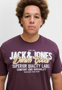 Tumedast puuvillast T-särk, millel on julge valge ja kollane tekst "JACK & JONES Denim Goods", lõdva istuvuse ja ümarate kaelustega.