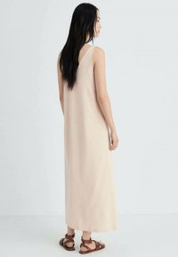 Lichtbeige mouwloze maxi-jurk met een relaxte pasvorm, gemaakt van zacht materiaal, met een ronde rug. Gekleed met bruine sandaaltjes met bandjes.