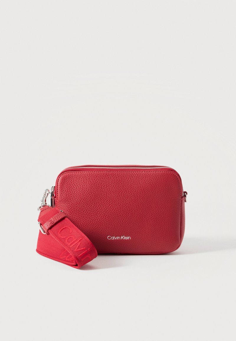 Κόκκινη τσάντα Calvin Klein από ανάγλυφο δέρμα με πλατύ ρυθμιζόμενο λουρί και ασημί φερμουάρ σε απλό λευκό φόντο.