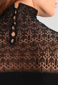 Svart spetsplagg med hög krage och invecklade mönster. Knäppning bak med fem runda svarta knappar. Mjuk textur.