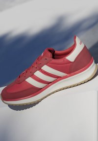 Chaussure de sport rouge avec des rayures blanches, semelle beige et lacets, positionnée sur une surface blanche avec un fond d'ombre bleu.