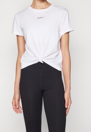 Mujer con camiseta blanca de manga corta anudada en la cintura y leggings negros ajustados sobre un fondo liso.