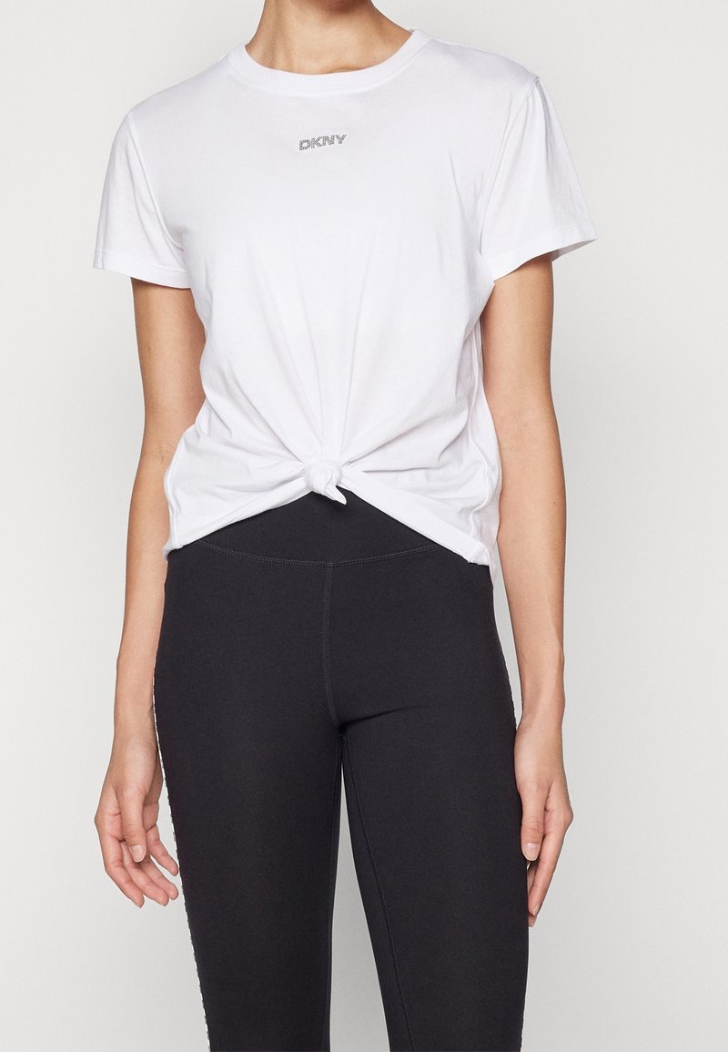Femme portant un t-shirt blanc à manches courtes noué à la taille et des leggings noirs ajustés sur un fond uni.