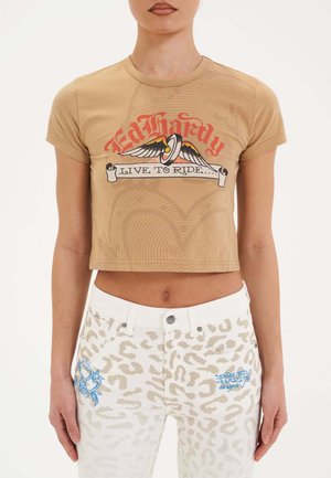 Beige, kurzes T-Shirt mit kurzen Ärmeln, das ein grafisches Design in Rot und Blau zeigt. Kombiniert mit weißen Jeans, die ein dezentes Leopardenmuster aufweisen.