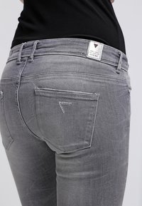 Vaqueros de denim gris con corte slim; cuentan con un logo triangular con gemas en el bolsillo trasero y una etiqueta de marca en la cinturilla.