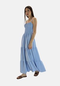 Blaues, gestuftes Maxikleid mit gerafftem Oberteil, Spaghetti-Trägern und fließender Textur. Das Model trägt braune Sandalen und steht vor einem hellen Hintergrund.