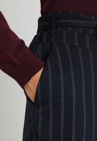 Granatowe spodnie w paski o dopasowanym kroju, z kontrastującymi białymi paskami i paskiem w talii. Widoczne ciemnoczerwone dzianinowe rękawy.