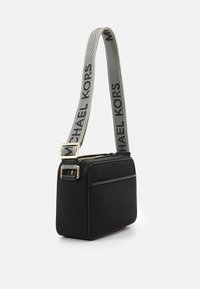Černá crossbody taška s texturovaným povrchem, zlatými kovovými detaily a šedým popruhem s tiskem "MICHAEL KORS" v �černé barvě. Obdélníkový tvar.
