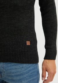 Black ribbed knit sweater with a textured surface and a brown leather logo patch near the hem. Fitted sleeves and a casual design.