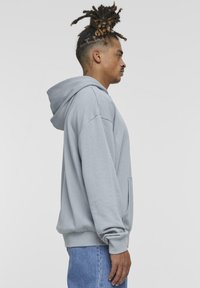 Gray hooded sweatshirt made of soft fabric, featuring a kangaroo pocket and ribbed cuffs. Model shown from the side, with denim jeans.