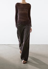 Haut à manches longues en maille côtelée de couleur grenat associé à un pantalon large à rayures grises. Sandales avec un accent circulaire visible. Fond uni.