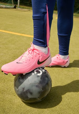 Nike Fußballschuhe in lebhaftem Pink mit schwarzen Akzenten, platziert auf einem schwarzen Fußball mit strukturierter Oberfläche, auf einem grünen Rasenfeld.