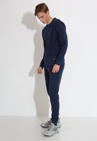 Tamno plava dukserica i hlače od pamuka, s opuštenim krojem, rebrastim mansetama i malim logom blizu rubova. Srebrne tenisice upotpunjuju izgled.