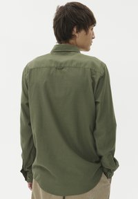 Zeleni dolg rokav s srajčnim ovratnikom, izdelan iz teksturirane tkanine, z gumbi na manšetah in ukrivljenim robom. Zadnji pogled.