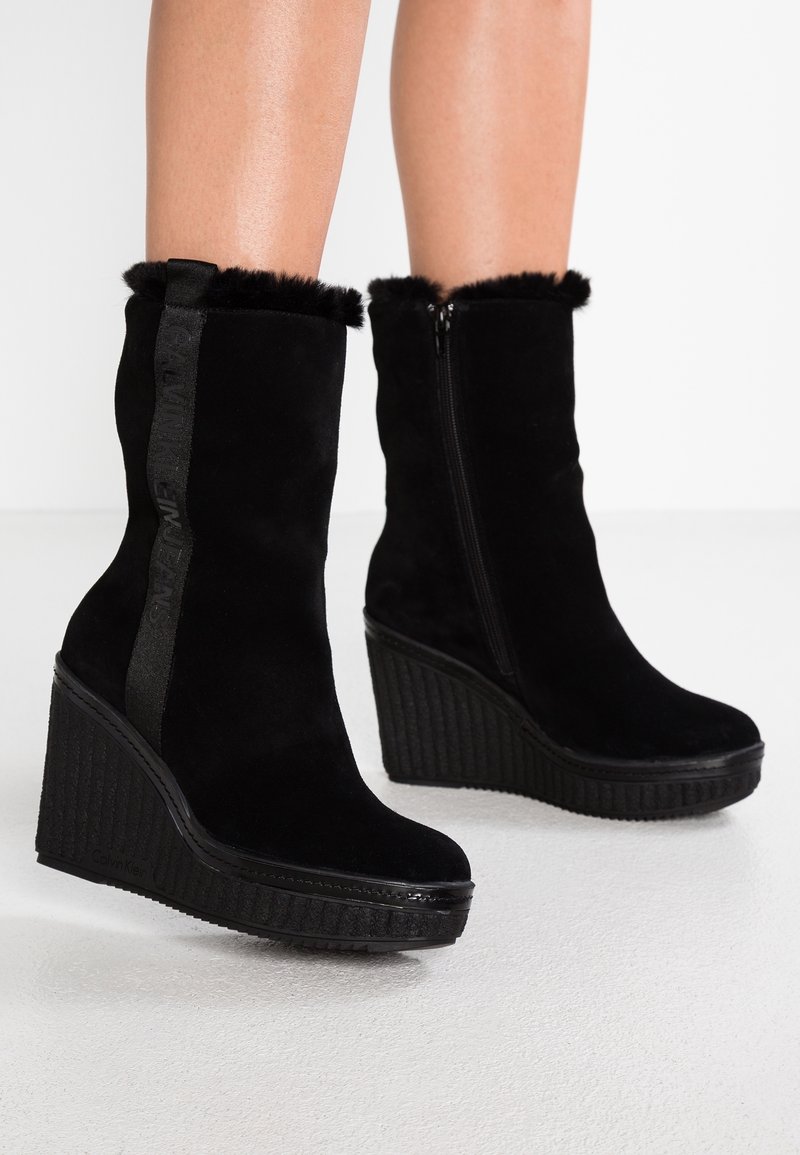 Bottines compensées noires avec bordure en fourrure, fermeture éclair latérale et bande verticale de marque, portées sur des jambes nues sur un fond blanc.