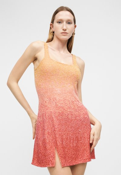 Γυναίκα που φοράει αμάνικο, πλεκτό φόρεμα με υφή σε διαβαθμισμένες αποχρώσεις από κίτρινο σε κοραλί με μπροστινή σχισμή, στέκεται μπροστά σε ένα απλό φωτεινό φόντο.