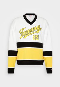 Maglia sportiva bianca con strisce orizzontali nere e gialle. Presenta "Tommy" in corsivo e "85" in grassetto giallo. Design a V con tessuto testurizzato.