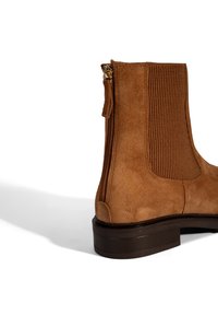 Bottine en suédine marron avec des panneaux latéraux élastiques côtelés et une fermeture éclair. Semelle épaisse de couleur marron foncé. Texture lisse avec une finition mate.
