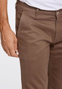 Braune Baumwollmischungshose mit glatter Textur, versehen mit einem Knopfverschluss, Gürtelschlaufen und Seitentaschen. Nahaufnahme des Stoffes.
