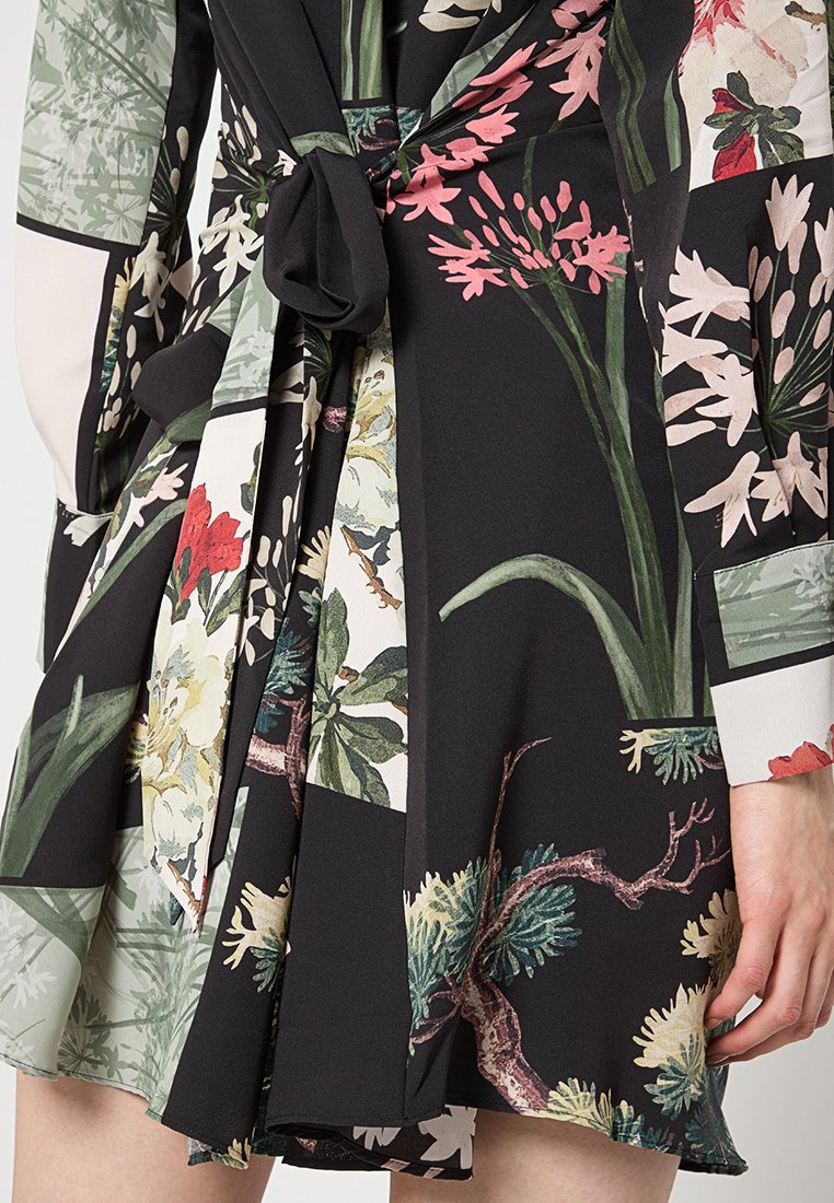 Zwart jurkje met bloemenpatroon, voorzien van een geknoopte taille, verschillende texturen en kleuren zoals roze, groen en wit, met opvallende botanische ontwerpen.