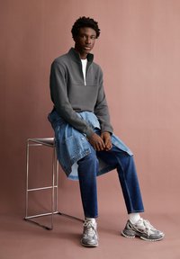 Sudadera gris con cremallera y cuello redondo blanco, combinada con una chaqueta de mezclilla azul, acompañada de jeans oscuros y zapatillas plateadas.