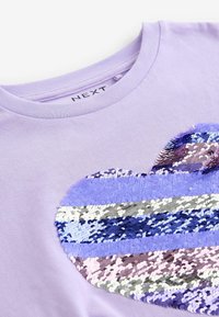 Lila T-Shirt mit kurzen Ärmeln und einem strukturierten Herzdesign aus Pailletten in verschiedenen Lila-, Silber- und Rosatönen. Etikett im Halsbereich sichtbar.