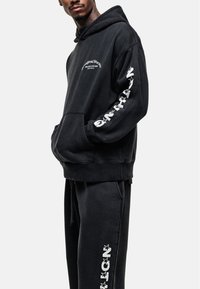 Zwarte hoodie en bijpassende sweatpants gemaakt van een katoenmix. Kenmerken zijn een voorzak, gedurfde witte letters op de mouwen en een koordsluiting in de taille.