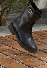 Bottine noire en cuir avec lacets et détail de boucle sur le côté. Talon étroit, semelle texturée et finition lisse. Positionnée au-dessus d'une surface texturée.