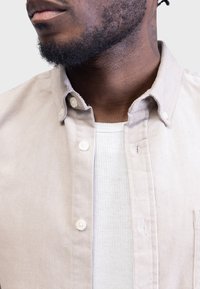Beige corduroy knap-skjorte med standard krave, der har en hvid ribbet undertrøje synlig under. Knapperne er runde og hvide.