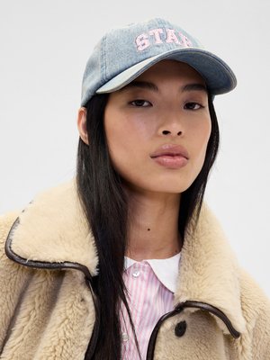 Lyseblå denimcap med "STA" broderi i rosa. Modellen har på seg en kremfarget faux fur-kåpe over en rosa og hvit stripete skjorte.
